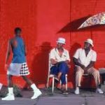 A screenshot from Spike Lee's Do the Right Thing. Four young Black men walk near three seated older Black men.